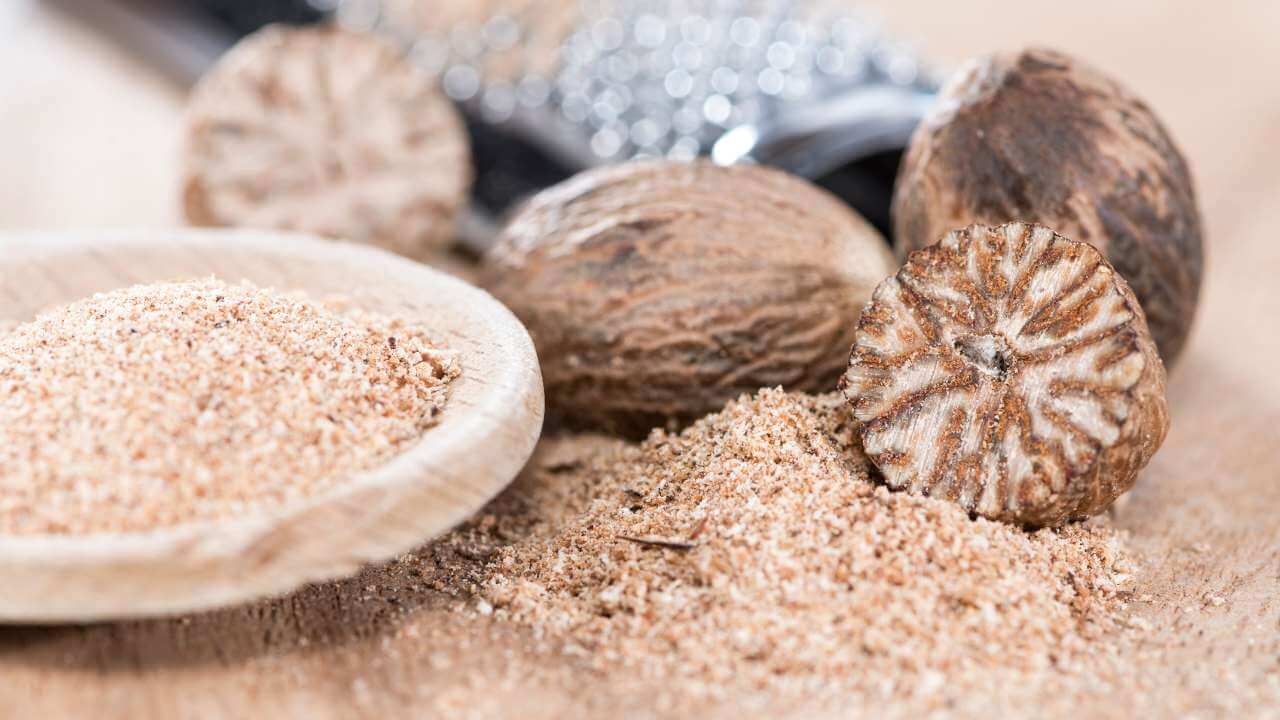 3 nutmeg seeds sitting on top of a small mound of nutmeg powder beside a wooden bowl with a grater blurred in background.  