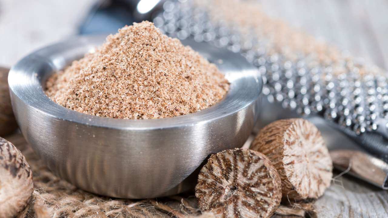 Nutmeg powder in a steel bowl beside a nutmeg seed with dark spots cut in half and a grater blurred in background.  