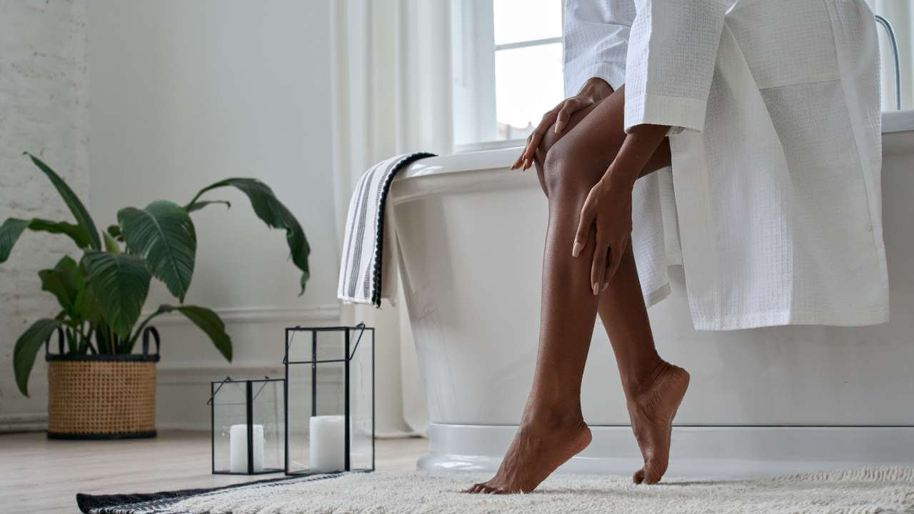 Woman with darker skin of color sitting on edge of bathtub in a white robe, assessing her smooth legs for eczema symptoms.