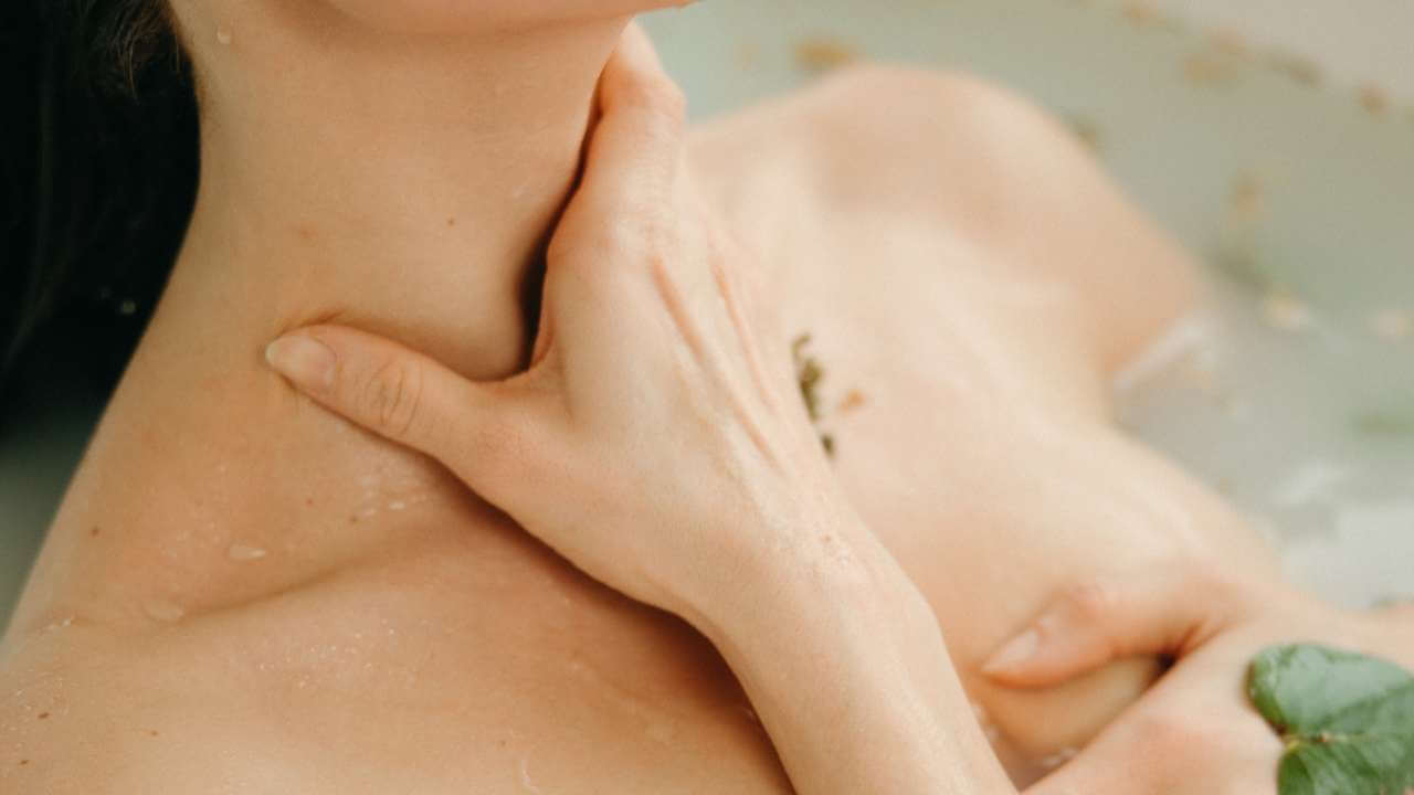 Closeup of a woman’s hand caressing her wet neck and chest while taking a spa bath full of botanicals. 