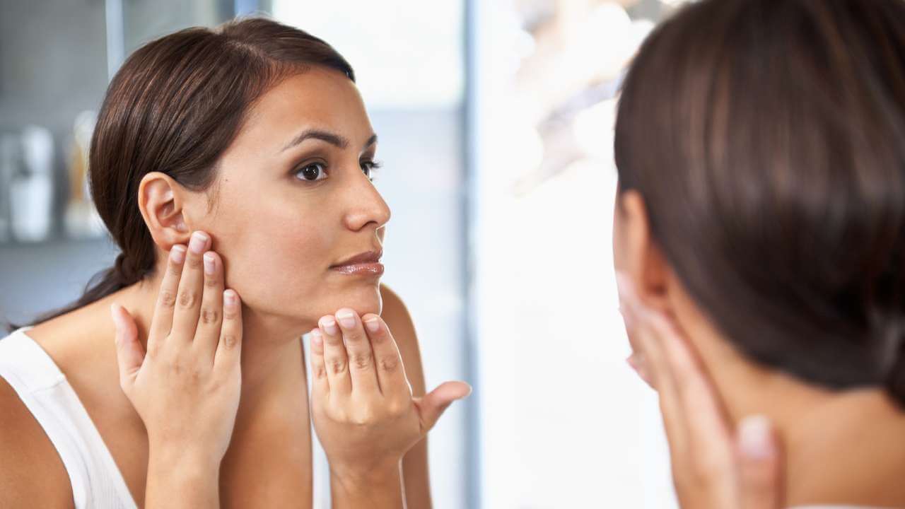 Closeup of a woman using both hands to examine the texture of the left side of her face while looking into a large mirror.