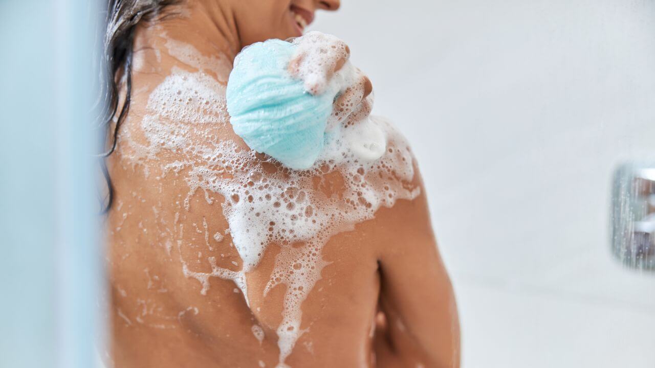 Closeup view of a happy woman washing the back of her right shoulder with soapy suds using a blue loofah sponge.  