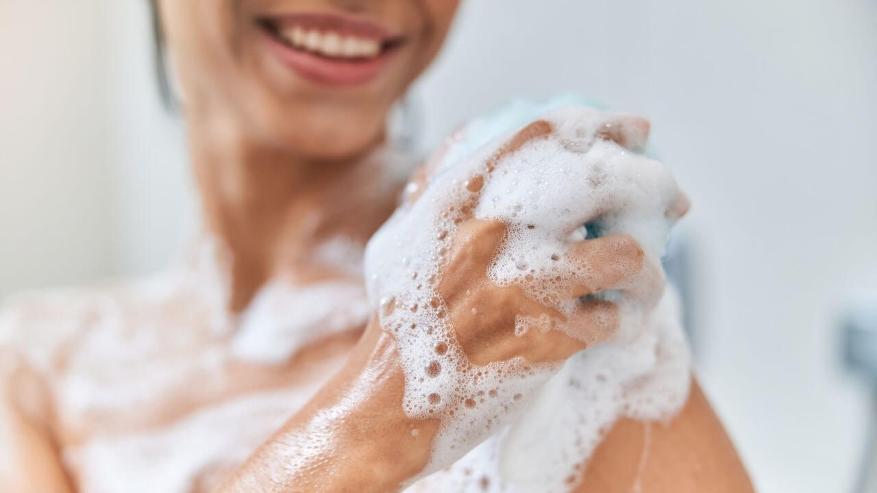 Closeup view of a smiling woman washing the front of her left shoulder with sudsy body wash using a blue loofah sponge.