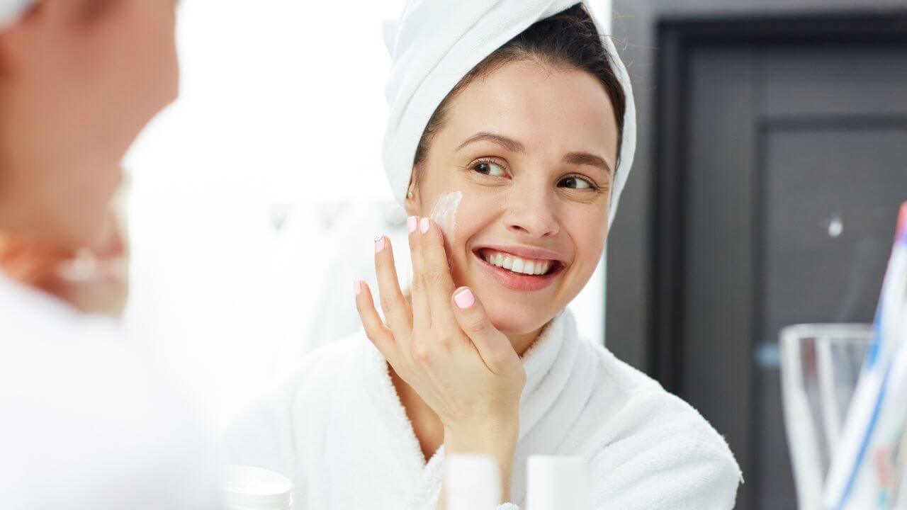 Smiling woman in white robe and hair wrapped in a white towel, looking in a mirror while using a non-comedogenic face cream. 