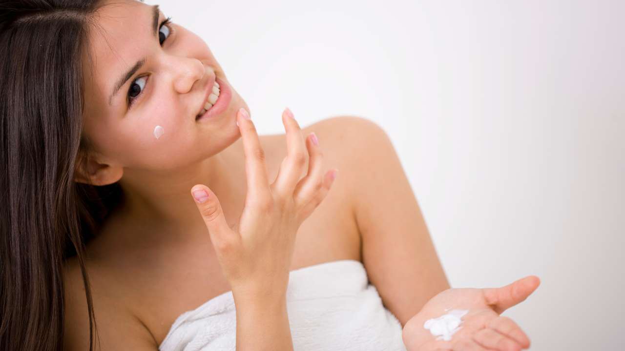 Smiling young woman in a towel with head tilted to the right, applying over the counter benzoyl peroxide cream to her chin.