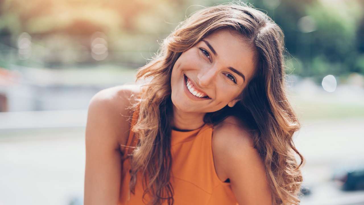 A smiling brunette woman wearing an orange sleeveless shirt, tilting her head to the left on a blurred outdoor background.