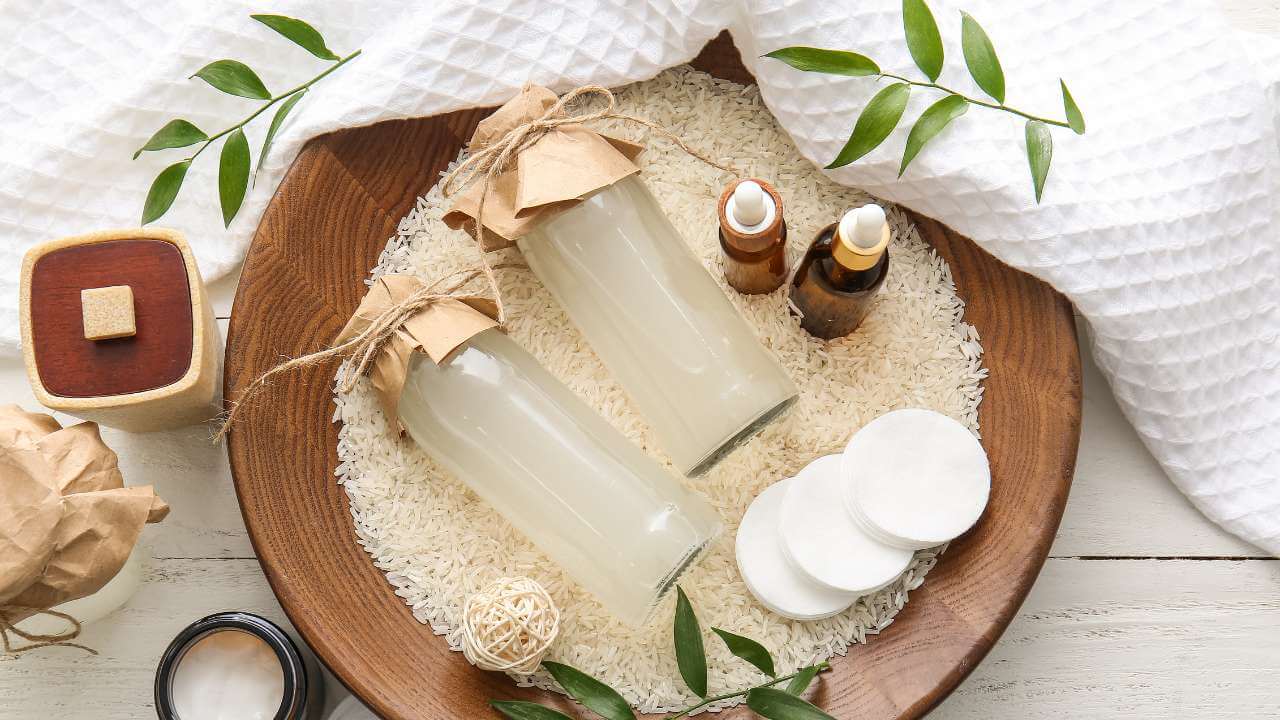 2 glass bottles of rice wine lying sidewise on a bed of rice on top of a wooden plate, bordered by skin care products.