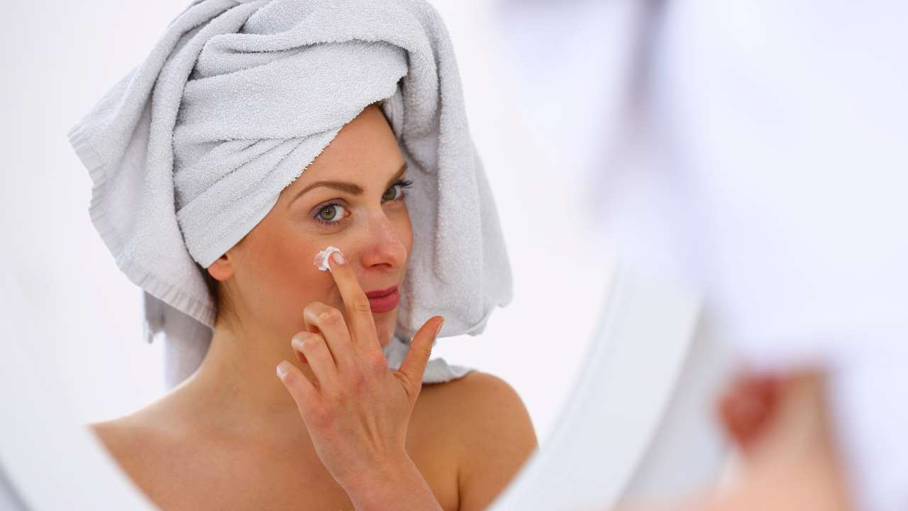 Woman with white towel wrapped around her head, applying moisturizing cream with ingredients like shea butter to her face.
