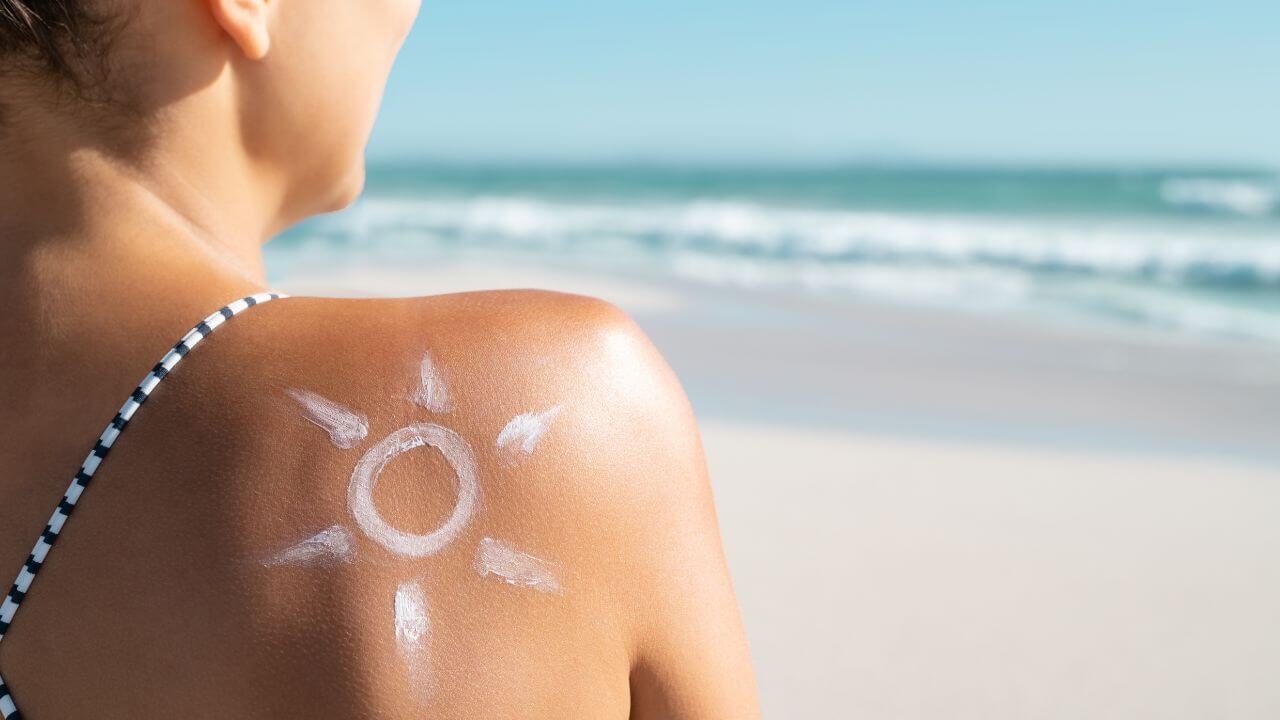 Sunscreen on a woman’s shoulder in the shape of the sun while sitting on the beach looking into the ocean in the background.
