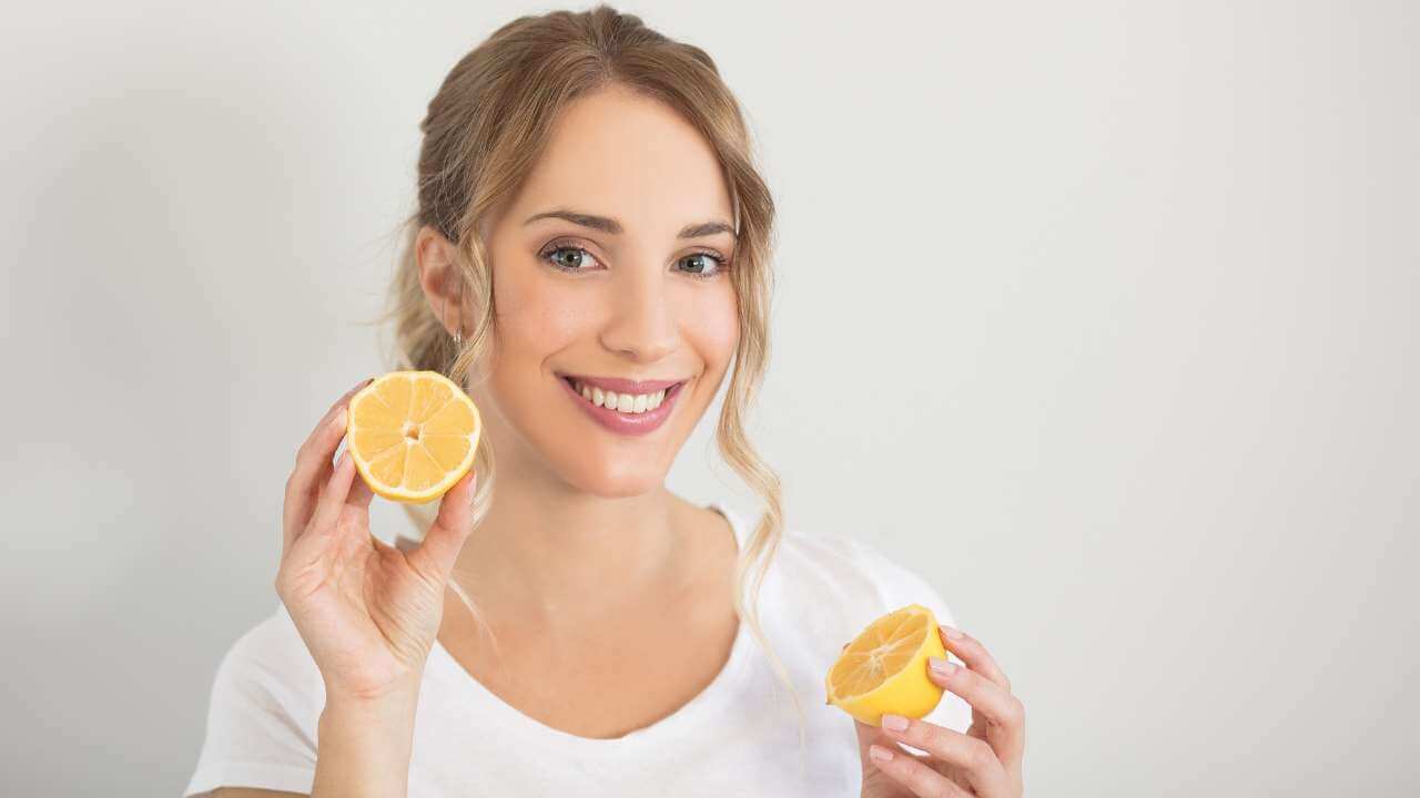 Pretty woman with a radiant glow holding half of a sliced lemon in each hand by her smiling face on a gray background.