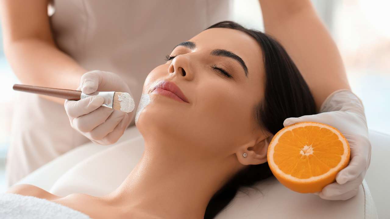 An esthetician holding an orange, uses a brush to apply a face mask clinically proven to reduce hyperpigmentation.
