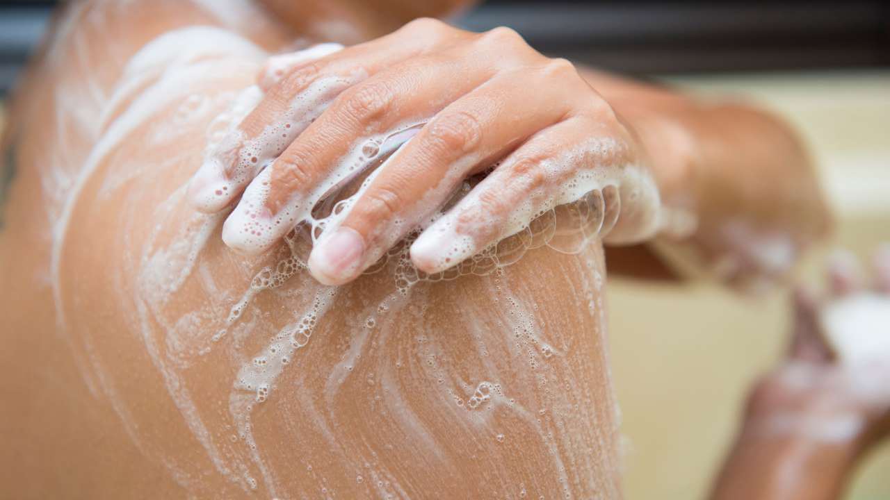 Closeup of a woman’s hand washing her shoulder with soapy suds while holding a bar of soap in the other hand during a shower.