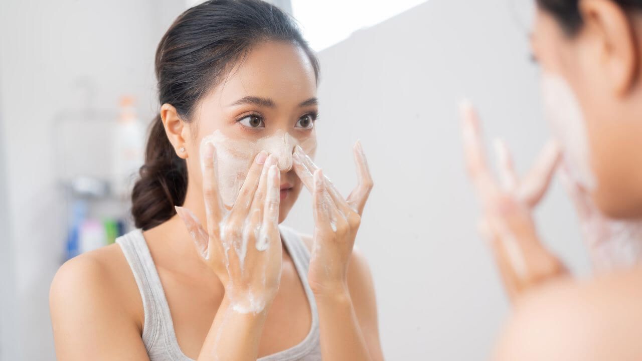 Asian woman looking into her bathroom mirror while washing her face at the sink to prevent age spots and other blemishes.