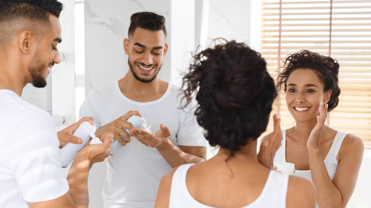 Couple smiling while washing their faces at bathroom sink, using a small amount of mild soap to help clear their adult acne. 