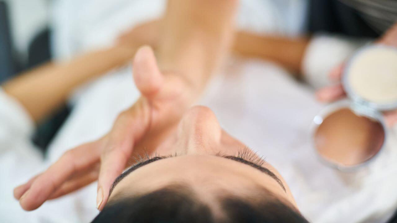 Top view of a woman lying comfortably on her back in a white robe while another woman’s hand applies primer to her left brow. 