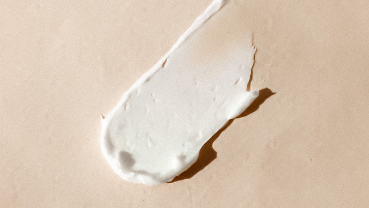 Close up of a thick smear of cream-colored primer, with the consistency of cake icing, on a peach-colored plain background.