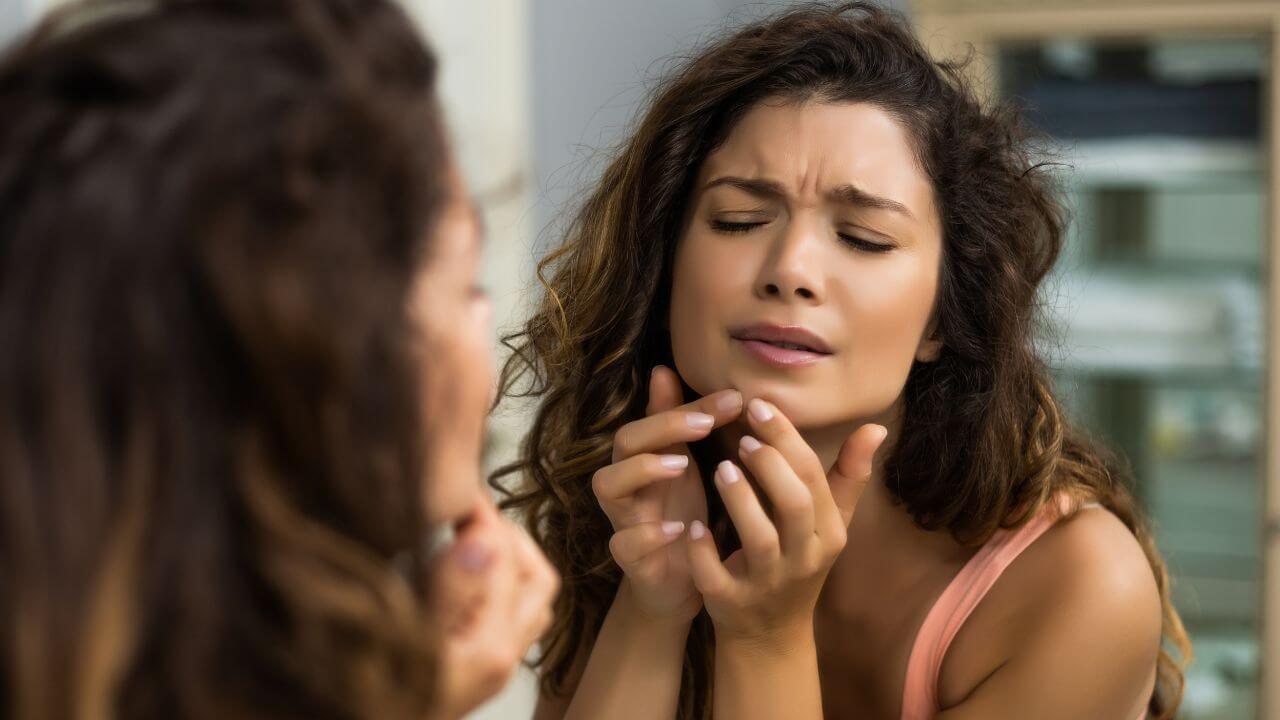 Close up of a young woman looking into a mirror with a painful expression, using both index fingers to pop a pimple on her chin.