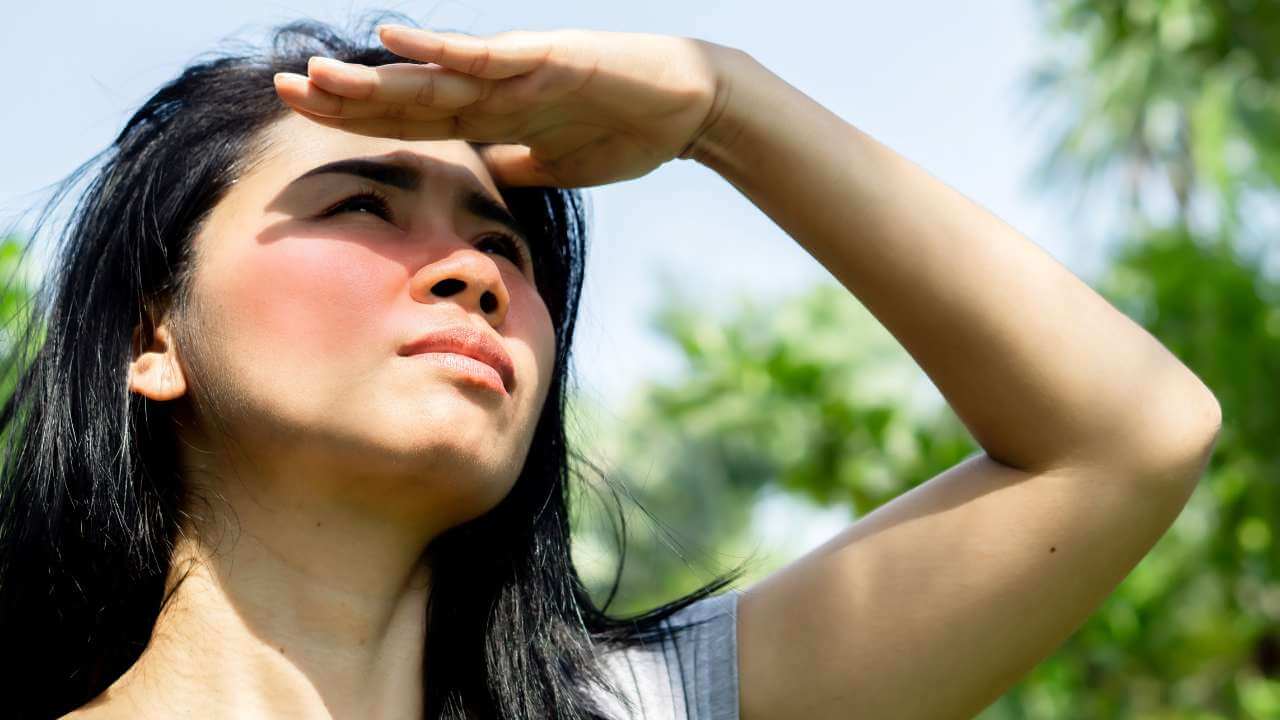 Asian female without sun protection, shielding her face with her hand to prevent sun spots while looking up at the sun.