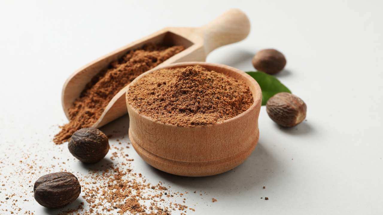 Wooden bowl and scooper full of nutmeg powder next to 4 nutmeg seeds against white background.