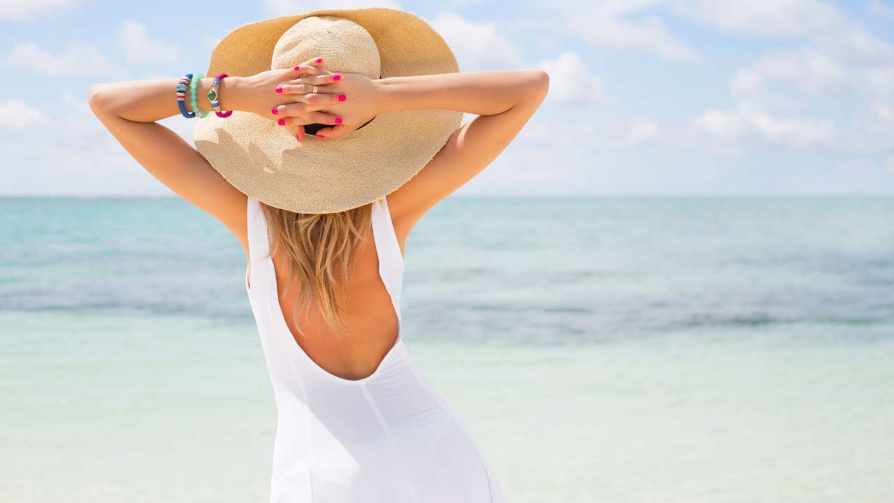 Back view of woman on beach, wearing an open back sundress and wide brimmed hat with hands behind her head facing the ocean.