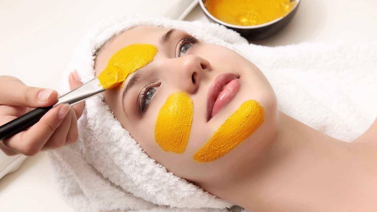 Esthetician using a brush to perform a patch test of a bright yellow turmeric paste to a young woman’s face. 