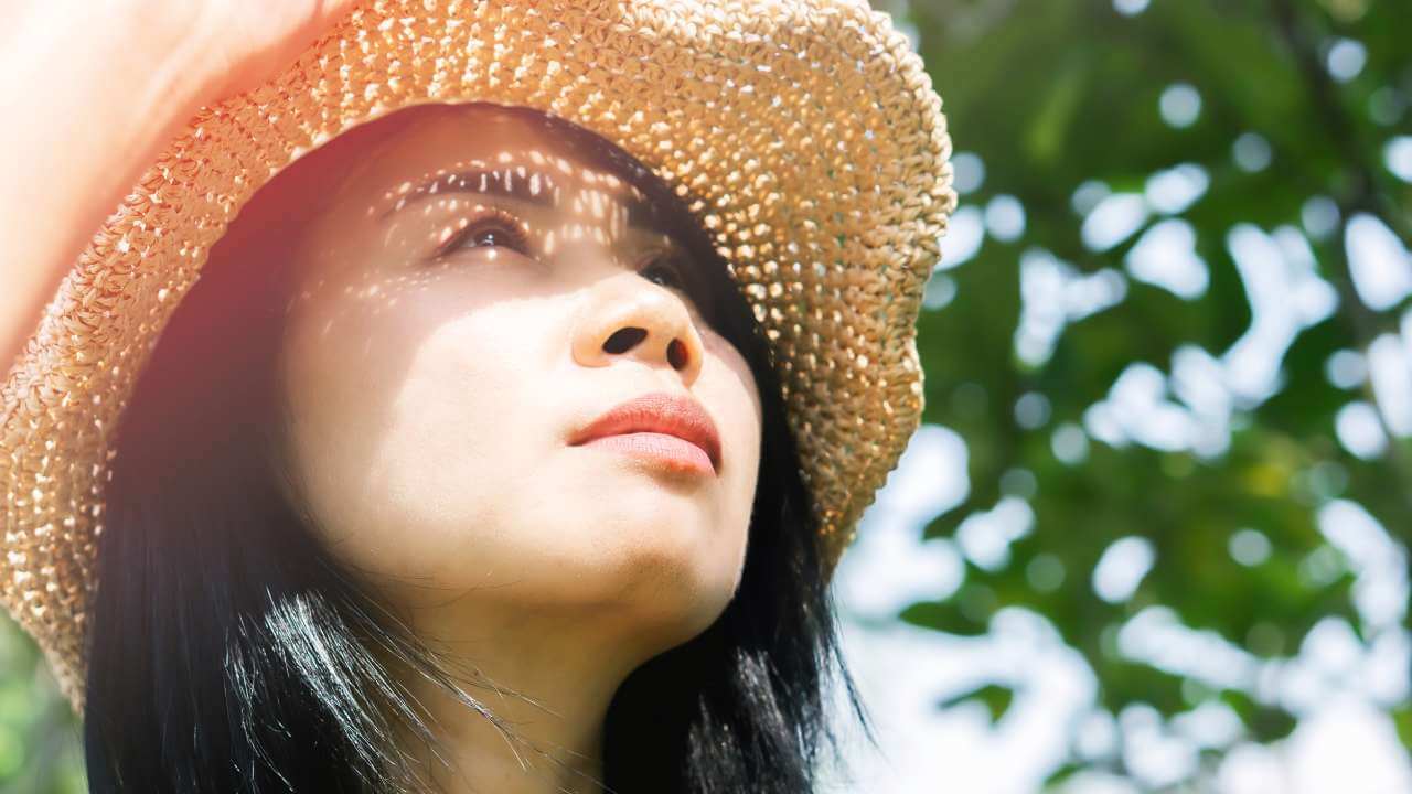 Asian woman outdoors looking up at the sky, shielding her face from high sun exposure by wearing a wide brimmed hat.